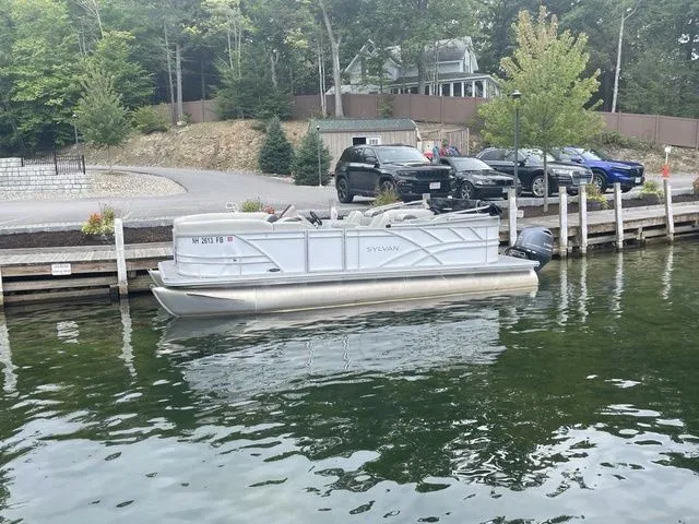 Slide: The Image of 2022 Sylvan 8522 LZ pontoon boat docked by a lakeside with parked cars nearby. - 1