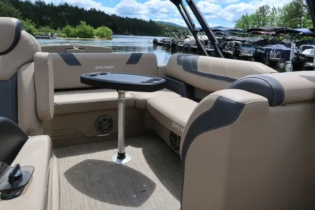 Slide: The Image of 2023 Sylvan L-3 DLZ BAR pontoon boat interior with beige seating and table, docked by a lake. - 7