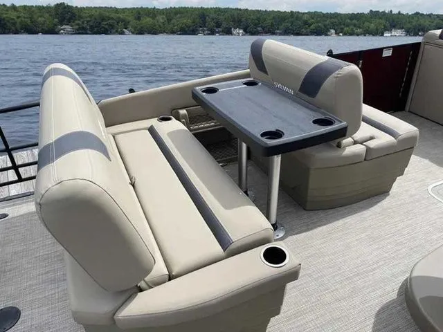 Slide: The Image of 2023 Sylvan L-1 DLZ pontoon boat interior with beige seating and table on a lake. - 5