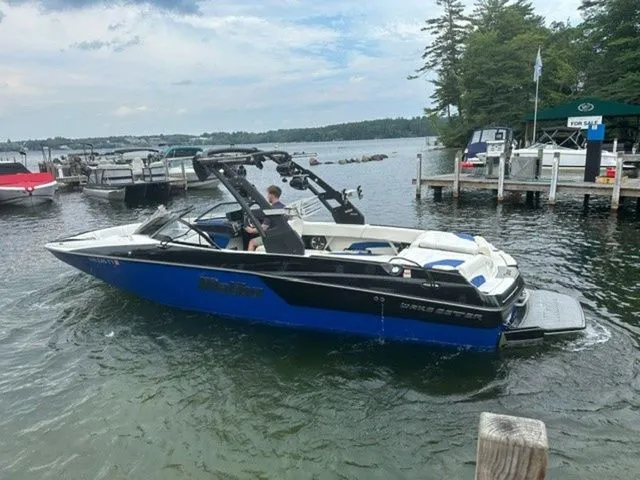Slide: The Image of 2017 Malibu 24 MXZ boat on a lake near a dock, surrounded by trees and other boats. - 20