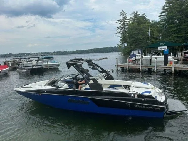 Slide: The Image of 2017 Malibu 24 MXZ boat on a lake near a dock with trees in the background. - 3