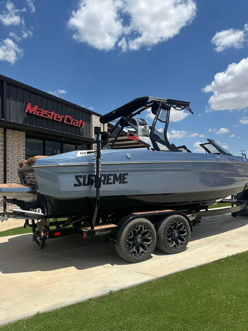 Slide: The Image of 2023 Supreme S220 boat on trailer outside MasterCraft building under blue sky. - 2
