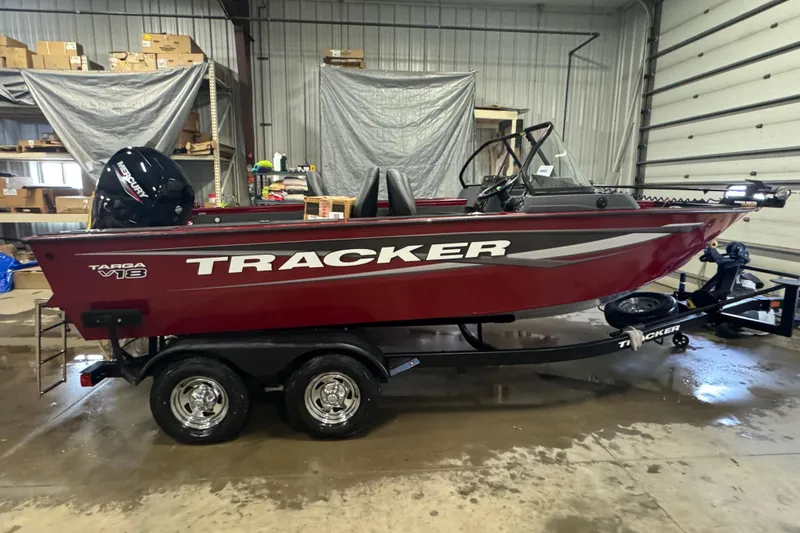 The Image of 2025 Tracker Targa V-18 WT boat in a garage, featuring a red hull and dual-axle trailer. - 0