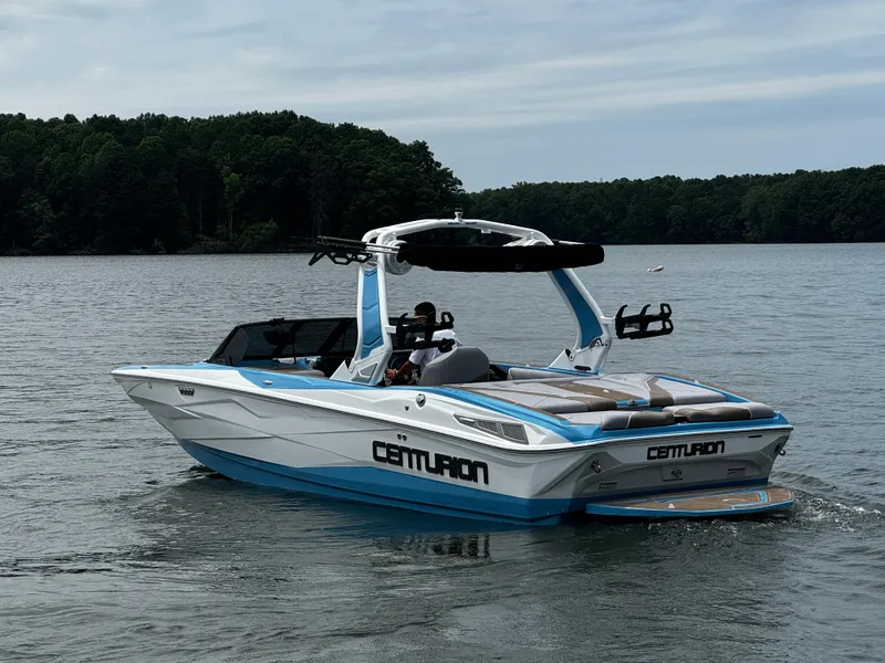 Slide: The Image of 2025 Centurion NV233 boat cruising on a lake with lush green forest backdrop. - 2