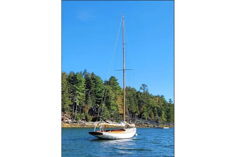 Slide: The Image of Sailboat Herreshoff Watch Hill 15, 1995 model, on serene water with forested backdrop. - 2
