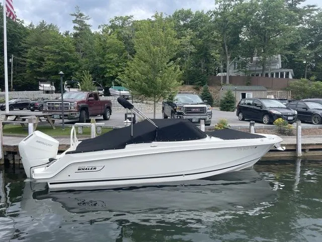 Slide: The Image of 2023 Boston Whaler 240 Vantage boat docked, covered, with trees and parked cars in the background. - 1