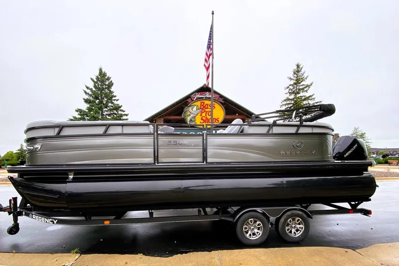 Slide: The Image of 2025 Regency 230 DL3 pontoon boat on trailer, displayed outside Bass Pro Shops. - 0