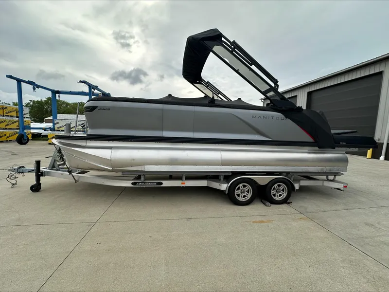 The Image of 2023 Manitou Explore pontoon boat on trailer, parked outdoors near a warehouse. - 0