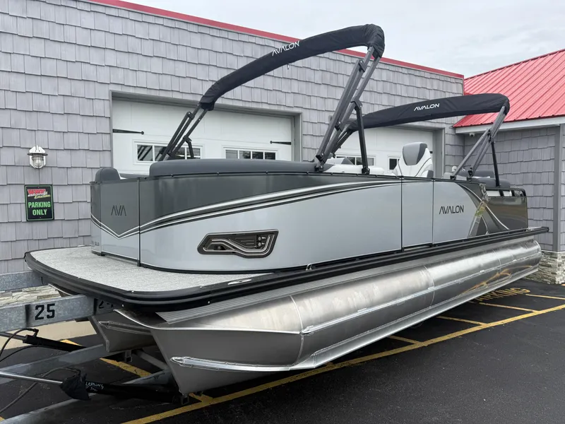 Slide: The Image of 2025 Avalon LSZ Quad Lounger 24' Tri Toon boat parked outside a building. - 2