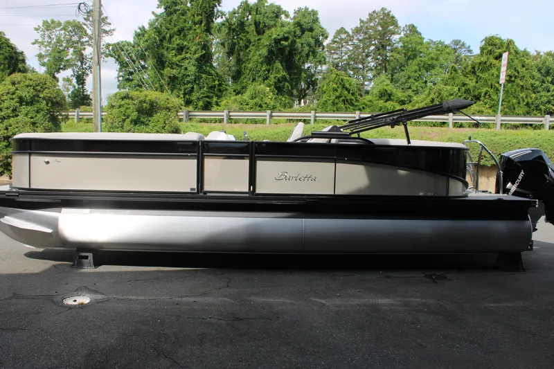 Slide: The Image of 2025 Barletta Aria 24QC pontoon boat parked on pavement, surrounded by greenery. - 12