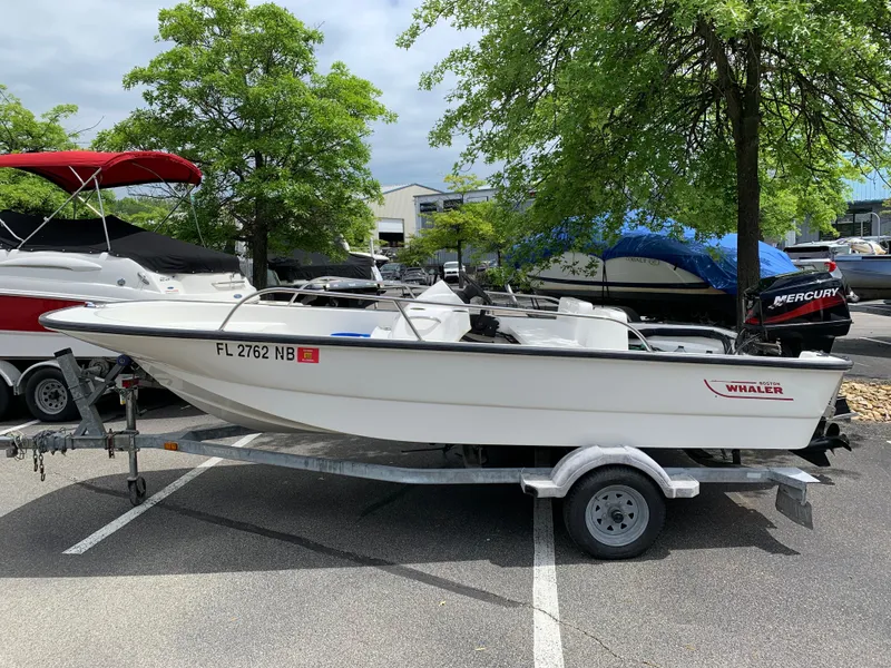 The Image of 2005 Boston Whaler 150 Sport boat on trailer in parking lot, surrounded by trees and other boats. - 0
