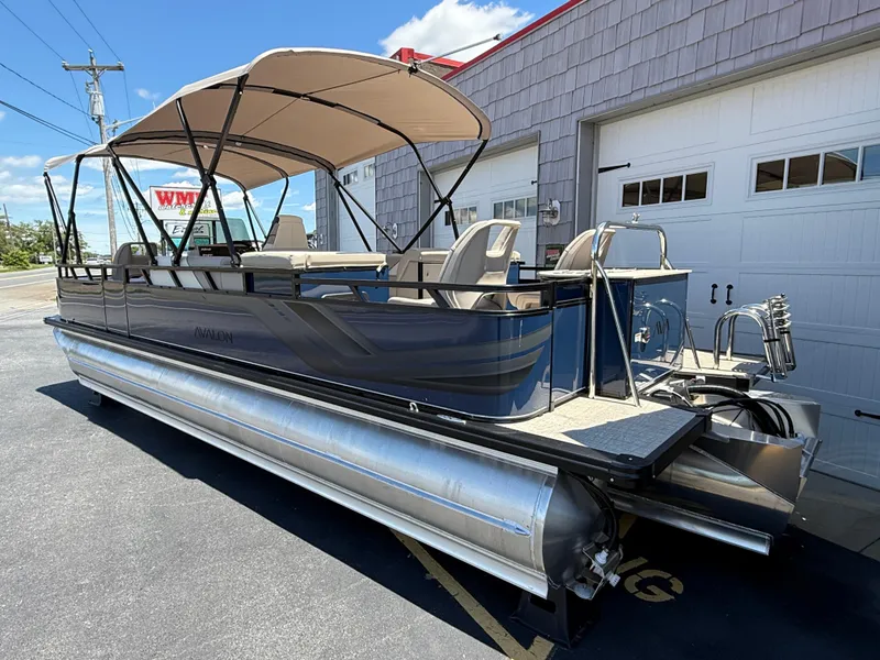 Slide: The Image of 2025 Avalon 2585 Venture 85 Tri Toon boat with center console, parked outside a garage. - 5