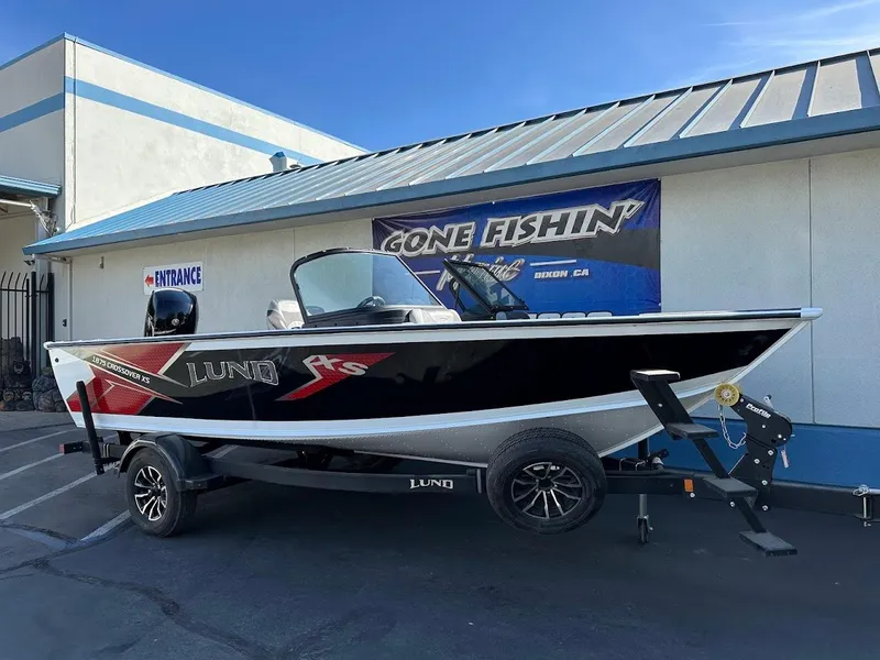 Slide: The Image of 2025 Lund 1875 Crossover XS boat on trailer, parked outside a building with "Gone Fishin'" sign. - 1