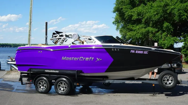 The Image of 2019 MasterCraft XStar boat in vibrant purple on a trailer by the water. - 0