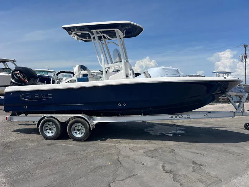 Slide: The Image of 2025 Robalo 226 Cayman boat on trailer, blue hull, parked outdoors under clear sky. - 2