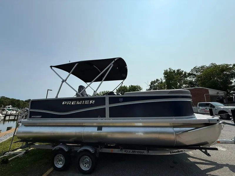 The Image of 2025 Premier 200 Sunscape RE XLC pontoon boat on trailer, black canopy, parked outdoors. - 1