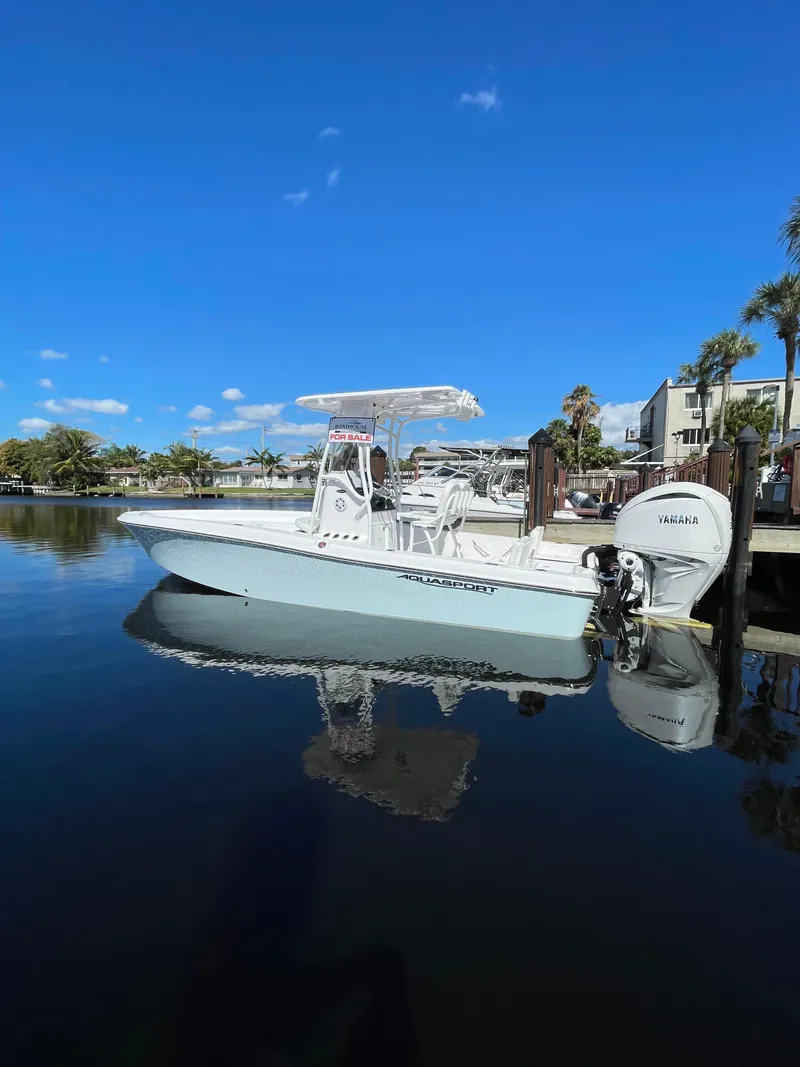Slide: The Image of 2024 Aquasport 230 Pro Bay boat docked on calm water under a clear blue sky. - 0
