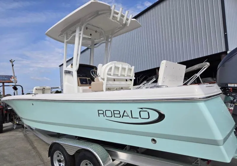 The Image of 2024 Robalo 246 Cayman boat on trailer, light blue hull, docked near a building. - 0