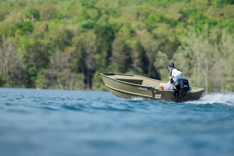 Slide: The Image of 2025 Lowe L1448M Jon boat on a lake, surrounded by lush green trees. - 6