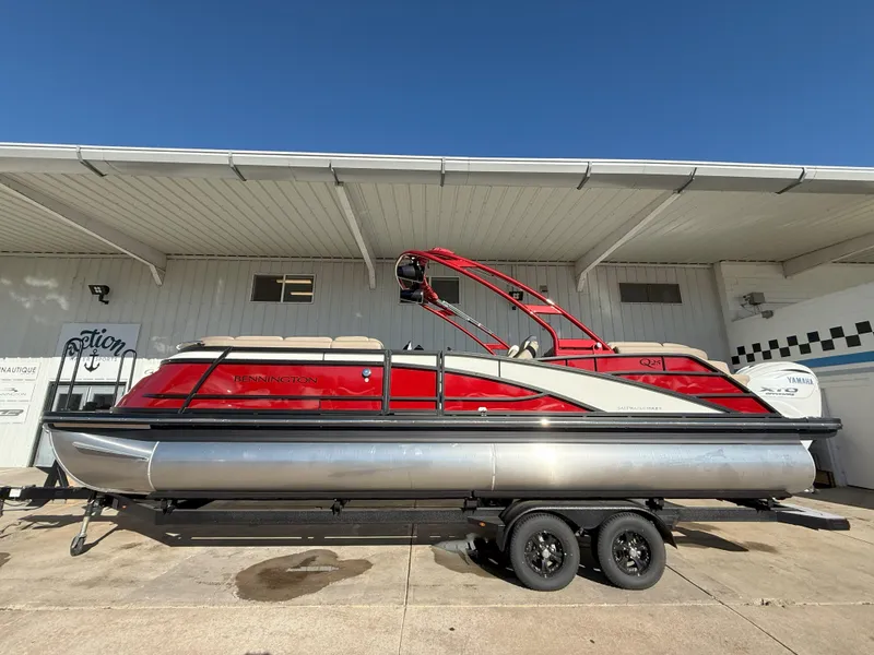 Slide: The Image of 2025 Bennington 25QXFB pontoon boat with red accents on a trailer, parked outdoors. - 0