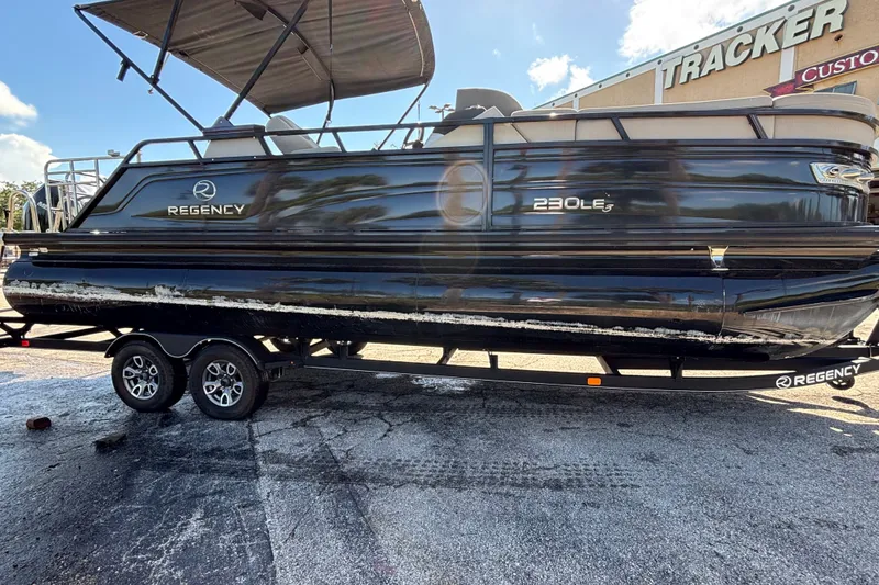 Slide: The Image of 2023 Regency 230 LE3 Sport pontoon boat on trailer, parked outdoors. - 2