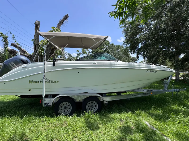 Slide: The Image of 2018 NauticStar DC 243 Sport Deck boat on trailer, parked on grass under a clear sky. - 6