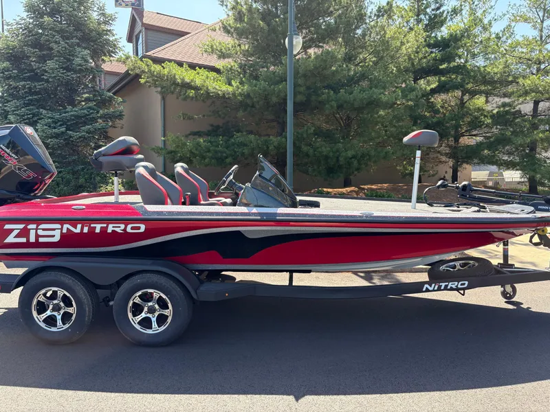 The Image of 2025 Nitro Z19 boat, red and black, parked on a trailer near trees. - 0