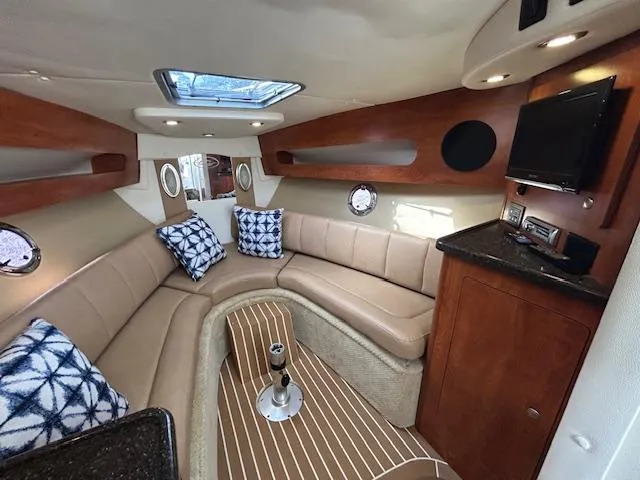 Slide: The Image of 2013 Rinker 290 Express Cruiser interior with beige seating, wood accents, and a mounted TV. - 15
