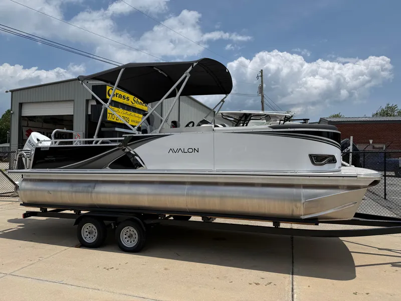 The Image of 2025 Avalon LSZ Versatile Rear Bench pontoon boat on trailer, parked outside under a cloudy sky. - 0