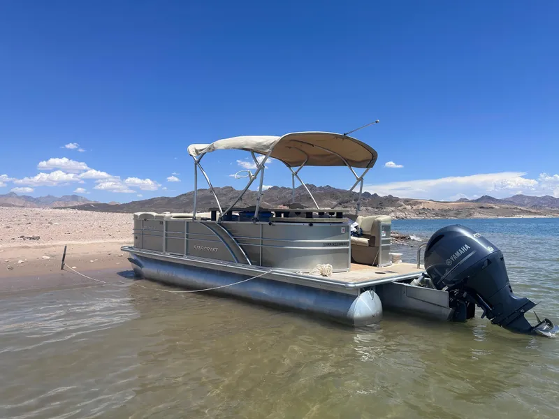 Slide: The Image of 2018 Starcraft 24 CXE Tritoon boat on sandy shore under clear blue sky. - 1