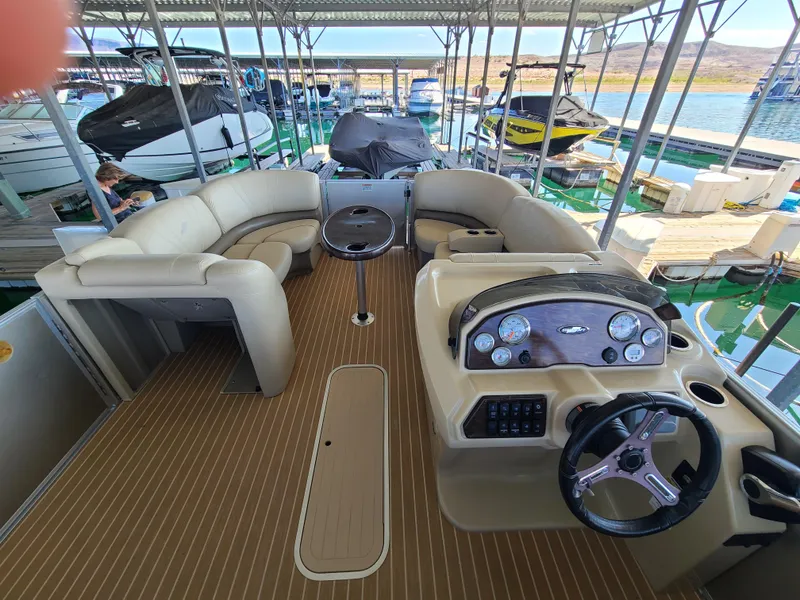 Slide: The Image of 2018 Starcraft 24 CXE Tritoon interior with beige seating and steering console in marina. - 6
