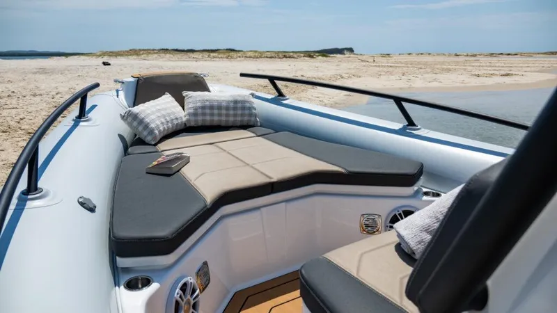 Slide: The Image of 2025 Grand Inflatables G750 boat with cushioned seating, pillows, and a book, on a sandy beach. - 9