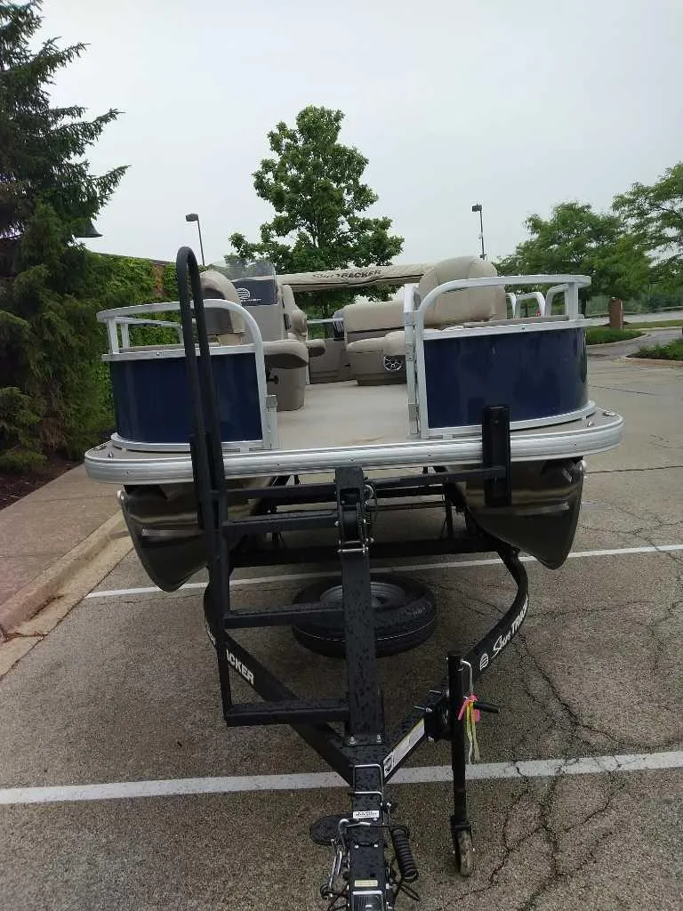 Slide: The Image of 2025 Sun Tracker Bass Buggy 18 DLX pontoon boat on trailer in parking lot. - 2