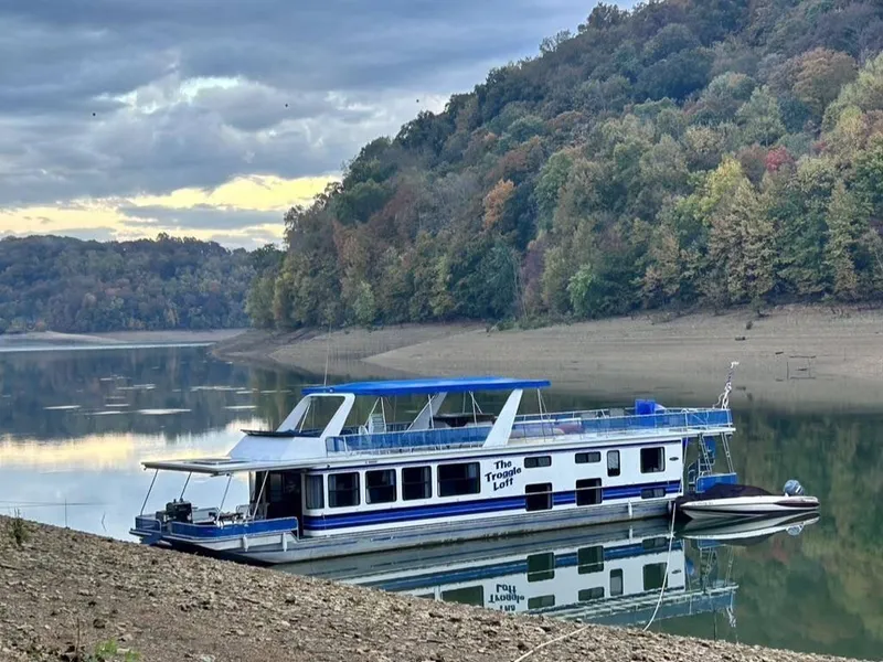 The Image of 1998 Stardust Cruisers 16x77 houseboat on a serene lake with forested hills. - 0