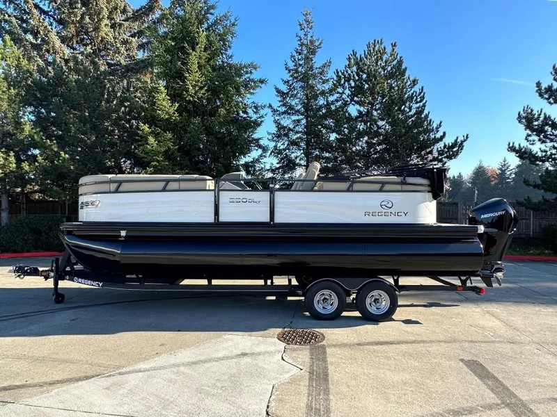 Slide: The Image of 2024 Regency 230 DL3 pontoon boat on trailer, parked outdoors with trees in background. - 0