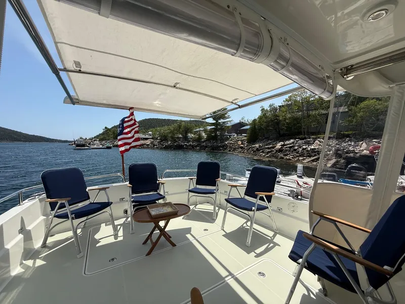Slide: The Image of 2019 Back Cove Downeast boat deck with chairs, table, and American flag, near rocky shoreline. - 17