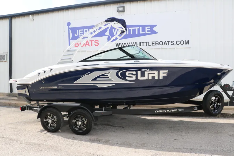 The Image of 2025 Chaparral 21 Surf boat on trailer, blue and white design, parked outside dealership. - 1