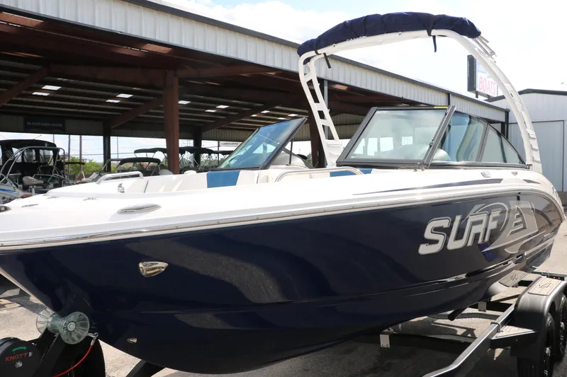 Slide: The Image of 2025 Chaparral 21 Surf boat with blue hull on a trailer, parked under a shelter. - 11