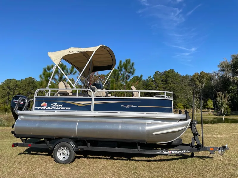 The Image of 2025 Sun Tracker Bass Buggy 16 XL Select pontoon boat on trailer, parked on grass. - 0