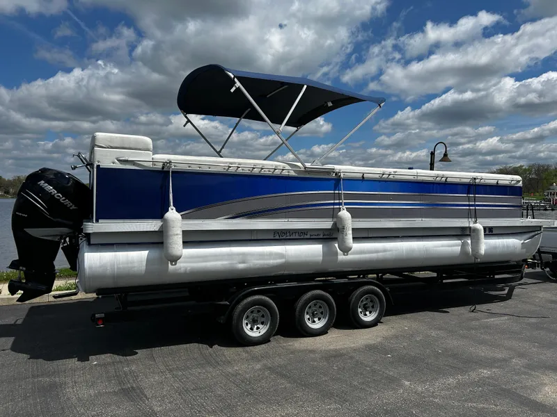 Slide: The Image of 2005 Evolution 26 pontoon boat with canopy on a trailer, parked by a lake under cloudy sky. - 14