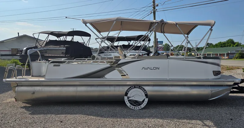 Slide: The Image of 2025 Avalon Platinum LSZ 2585 Versatile Rear Bench pontoon boat with canopy, parked on gravel. - 2