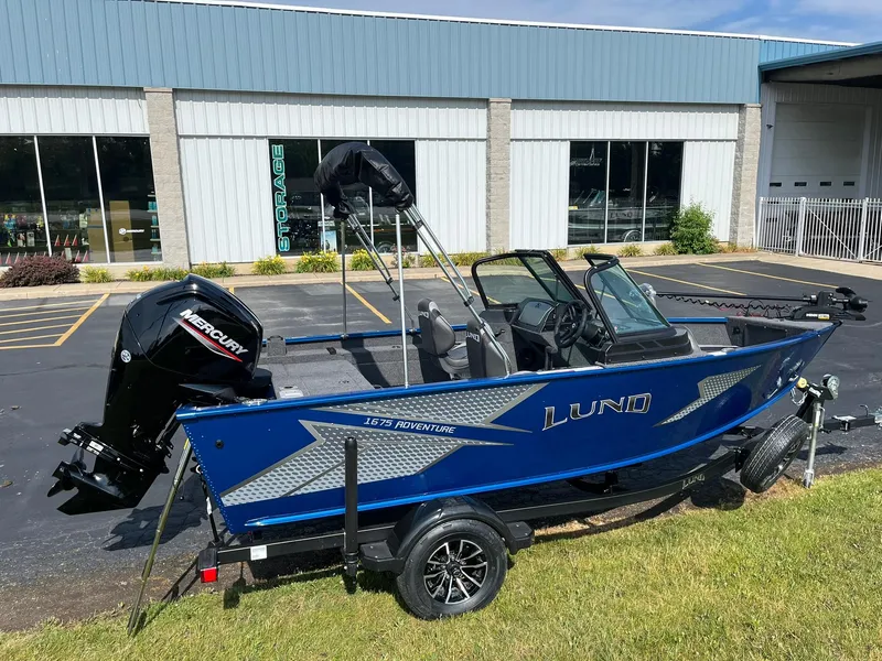 Slide: The Image of 2026 Lund 1675 Adventure Sport boat with Mercury engine, parked outside a building. - 2