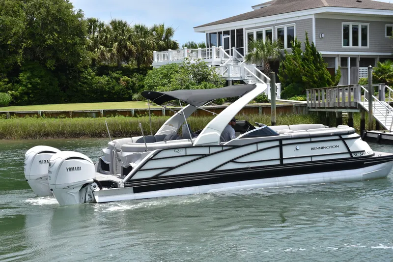 Slide: The Image of 2024 Bennington 27 QFB pontoon boat cruising near a waterfront home. - 17
