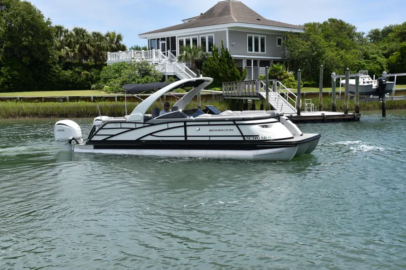 Slide: The Image of 2024 Bennington 27 QFB pontoon boat cruising near a waterfront house. - 18