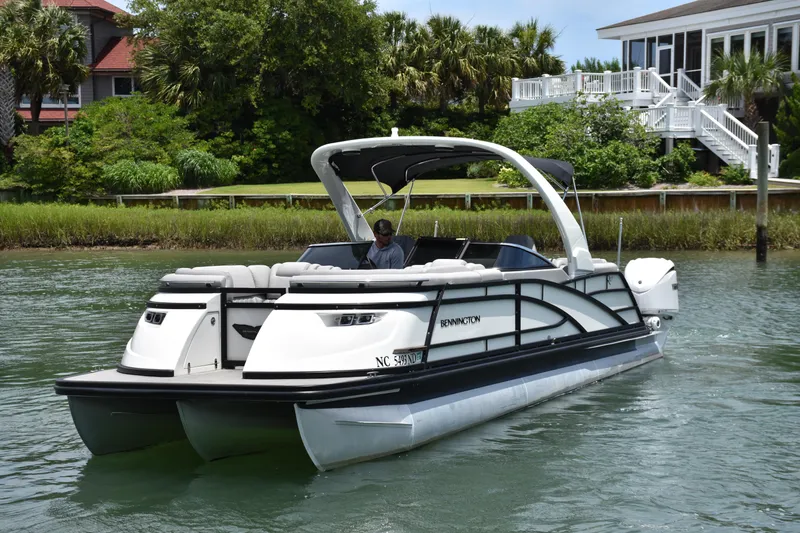 Slide: The Image of 2024 Bennington 27 QFB pontoon boat cruising on a scenic waterway near waterfront homes. - 27
