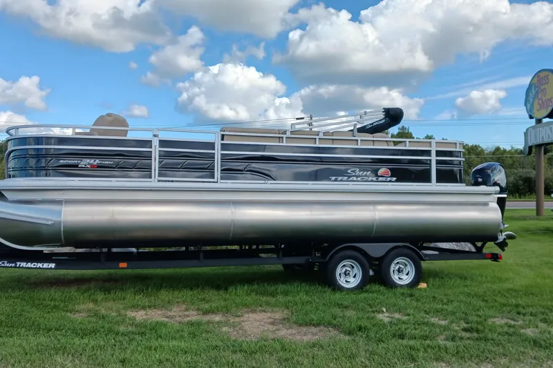 The Image of 2025 Sun Tracker Sportfish 24 XP3 pontoon boat on trailer, parked on grass under blue sky. - 0