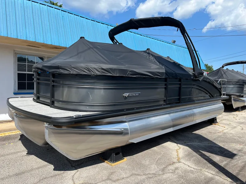 The Image of 2026 Harris Solstice 230 pontoon boat with black cover, parked outdoors under blue sky. - 0