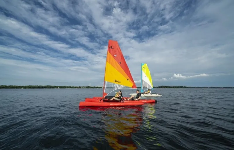 Slide: The Image of 2025 Hobie Mirage Tandem Island sailing on a calm lake under a partly cloudy sky. - 0