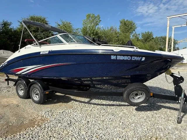 The Image of 2015 Yamaha SX210 boat on trailer, blue hull, parked on gravel. - 0