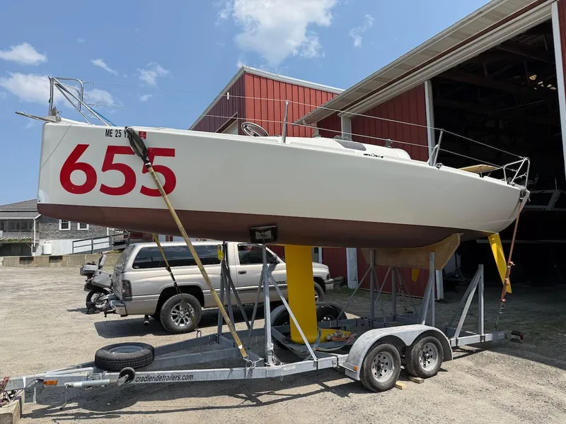 The Image of 2006 Mas Mini Transat 6.50 sailboat on trailer, parked outside a red building. - 1